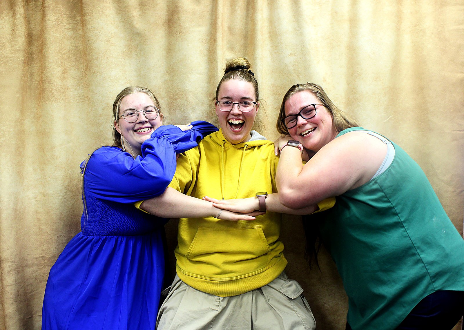 Three friends in blue, yellow and green leaning into a group hug, laughing, against a gold drape