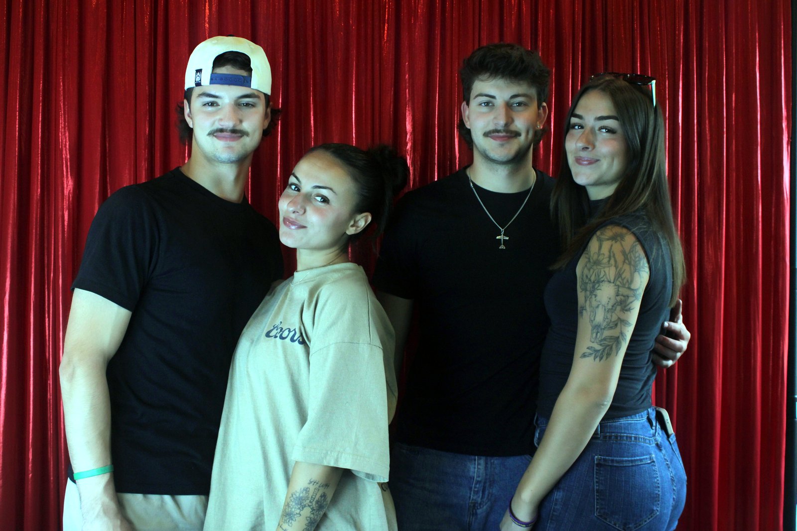 Four friends in black, one with a tattooed arm, standing shoulder to shoulder in front of the red curtain