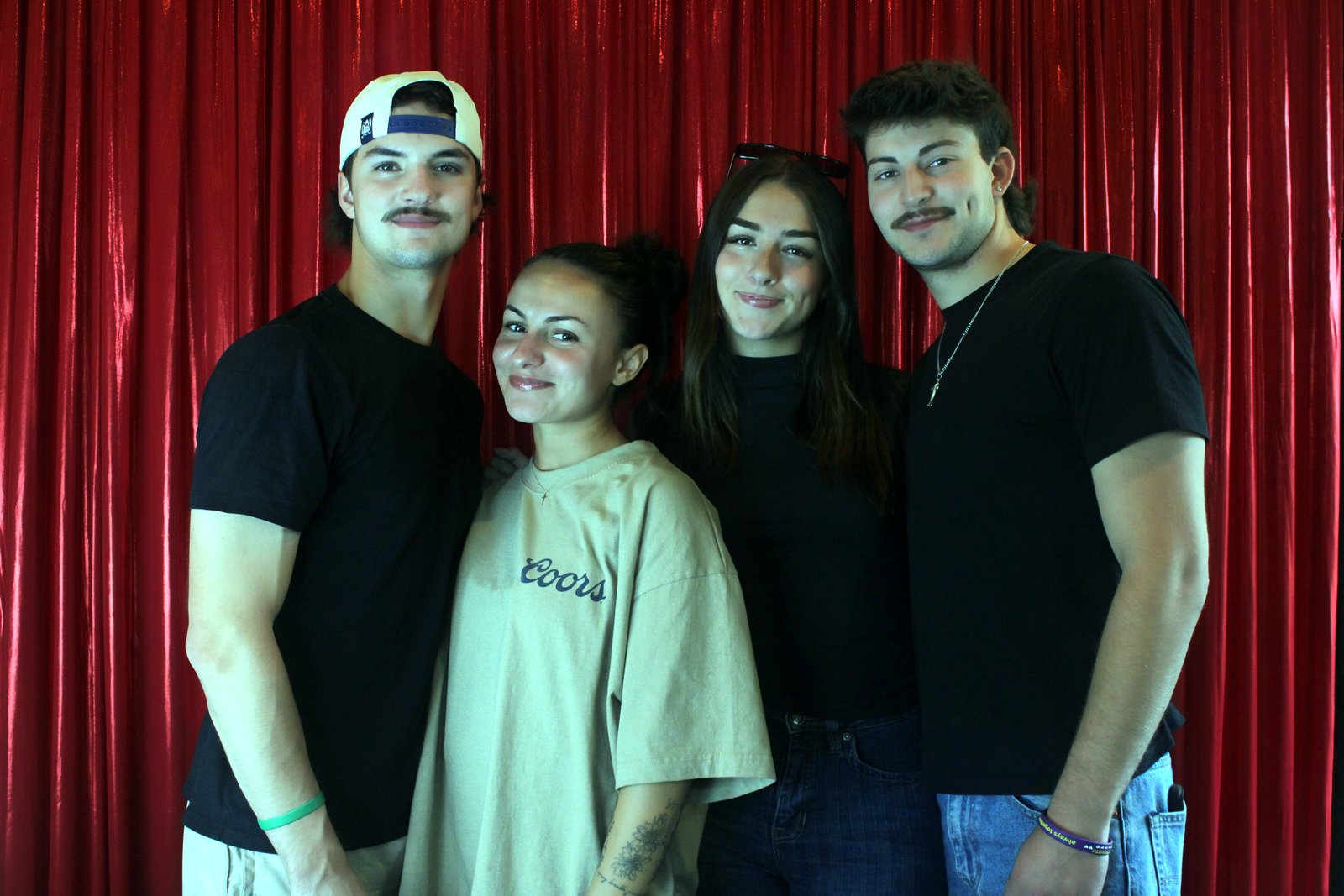 Four friends in black T-shirts and a Coors tee, arms around each other in front of the red curtain