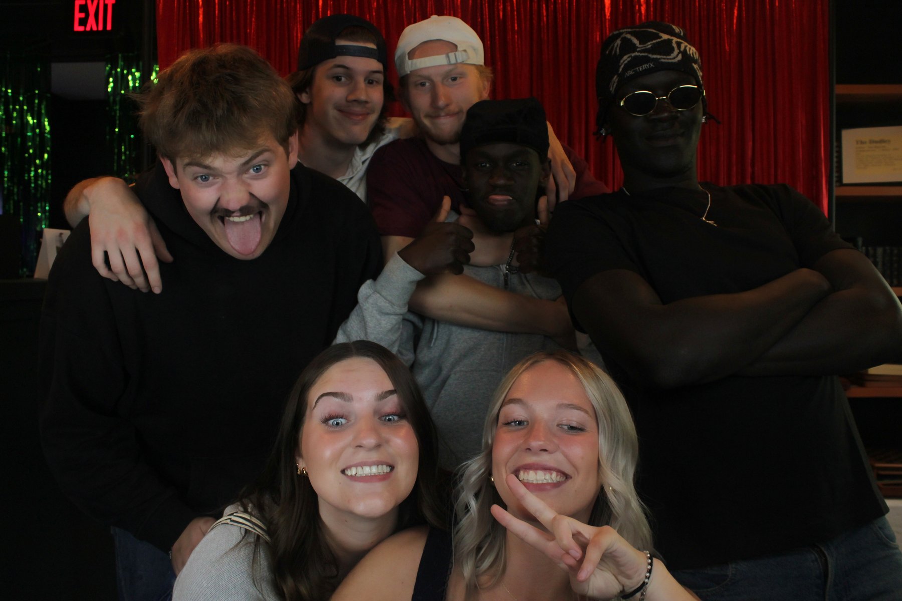 Six guests crowded in front of the red curtain backdrop, pulling faces and grinning at the mirror photo booth during a business's grand opening at The Dudley