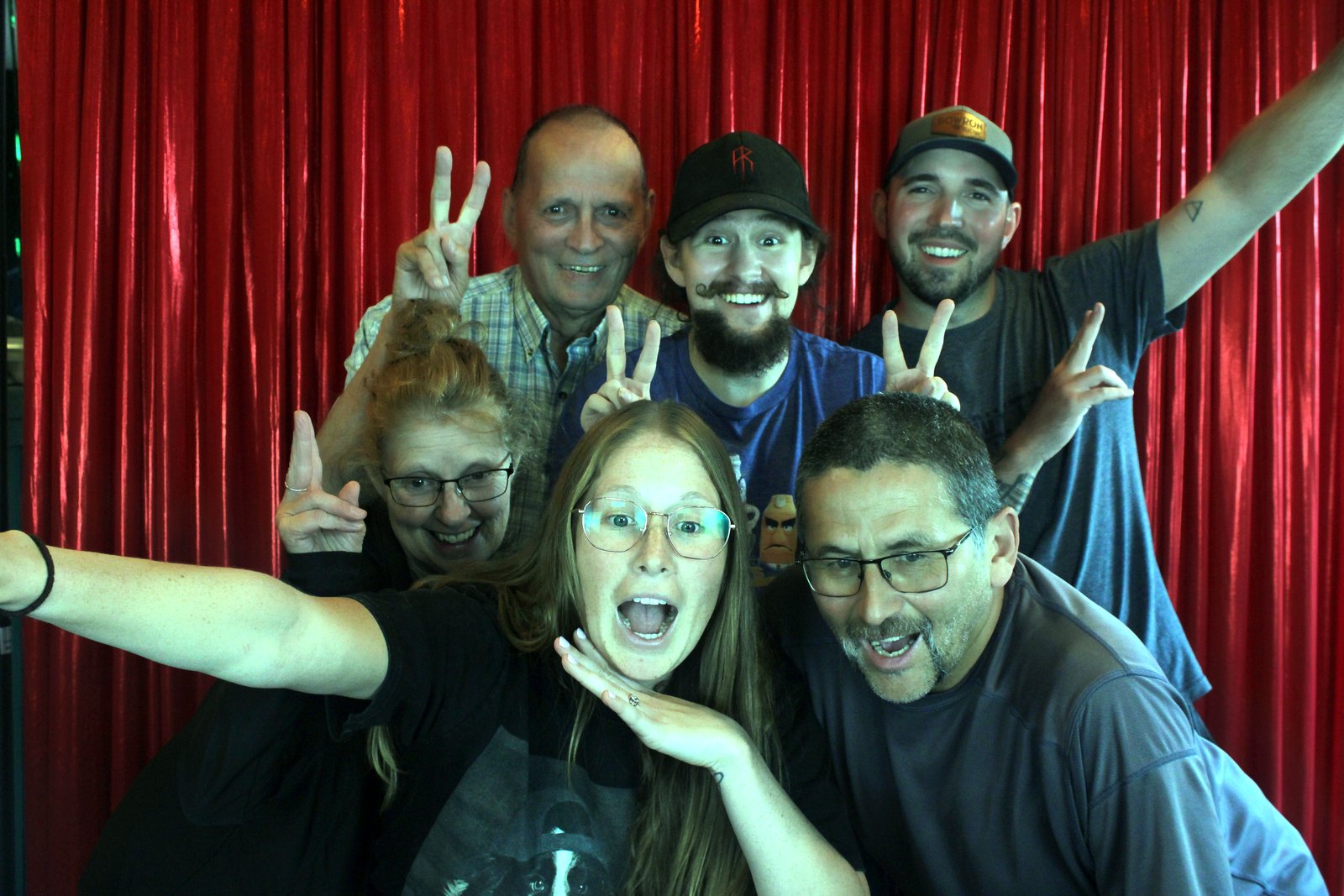 Seven people throwing peace signs and open-mouthed grins at the mirror booth, one arm stretched high into the frame