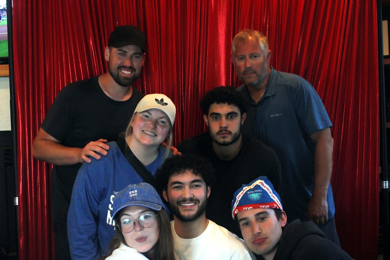 A trivia team of seven in ball caps and a blue toque grinning in two rows in front of the red curtain
