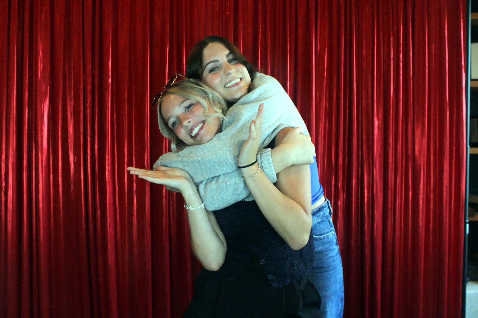 Two young women in a playful back-to-back pose, one in a grey cardigan and blue top, in front of the red curtain