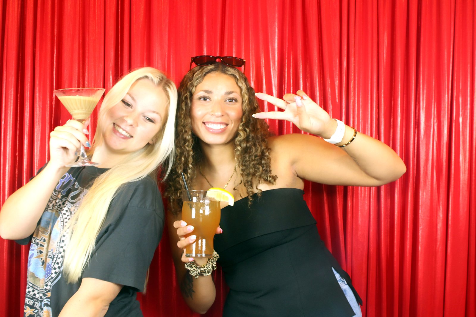 Two women raising a martini and a cocktail to the mirror booth in front of the red curtain