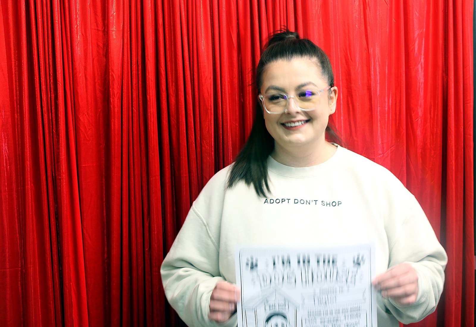 A woman in an Adopt Don't Shop sweatshirt laughing with her Dog House sign in front of the red curtain