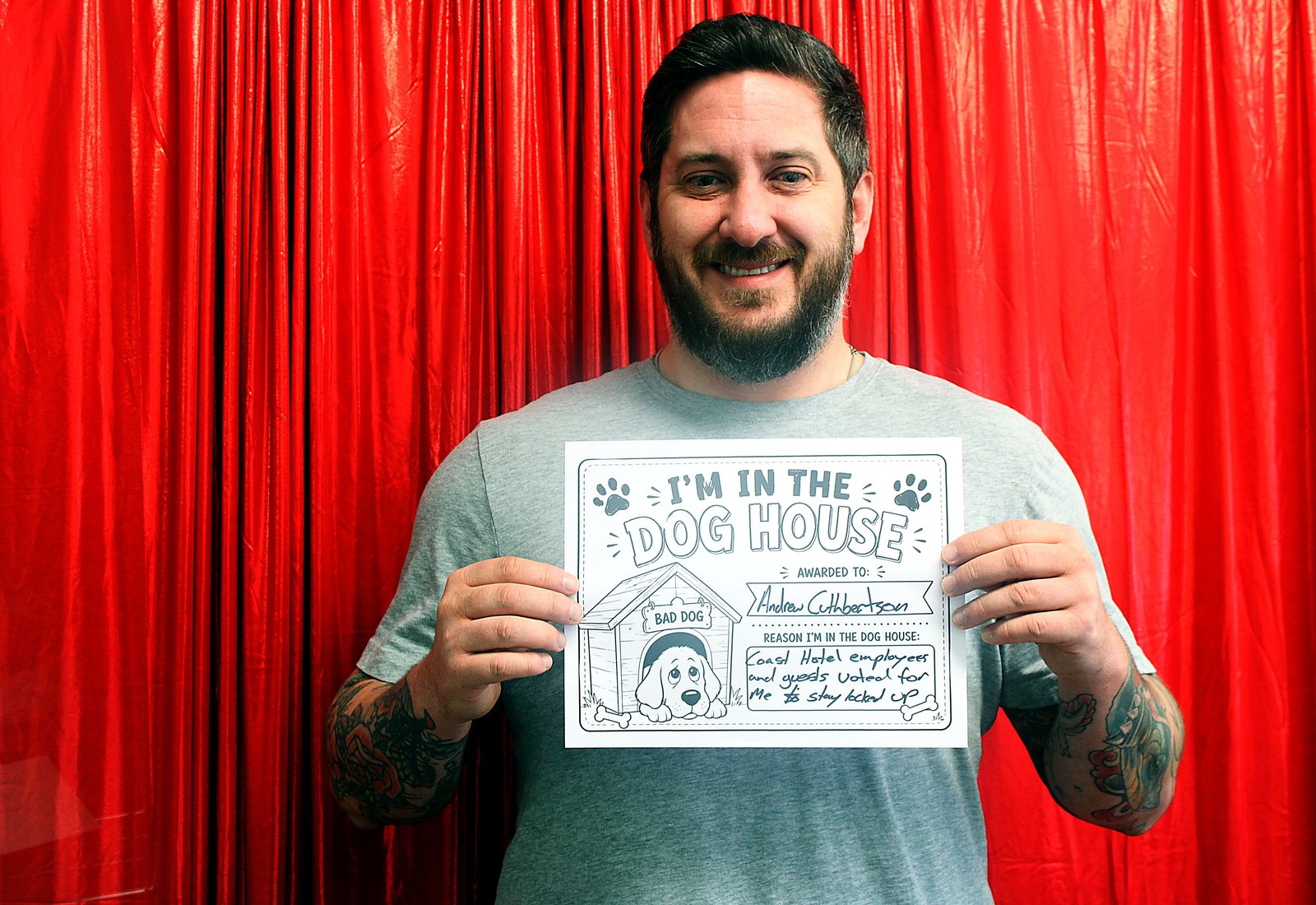 A smiling man with tattooed forearms holding an I'm in the Dog House sign in front of a red curtain